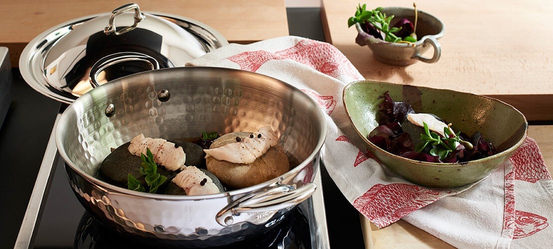 Edelstahl Kochtopf mit Deckel auf Induktionskochfeld, darin pochierter Fisch auf heißen Steinen, nebenbei eine Schüssel mit Salat und passendem Geschirrtuch Kochtopf aus Edelstahl auf einem Herd, darin drei Steine mit darauf serviertem weißem Fischfilet und frischen Kräutern, daneben eine grüne Schüssel mit Salat und einem weiteren Stück Fisch auf einem rot-weißen Geschirrtuch