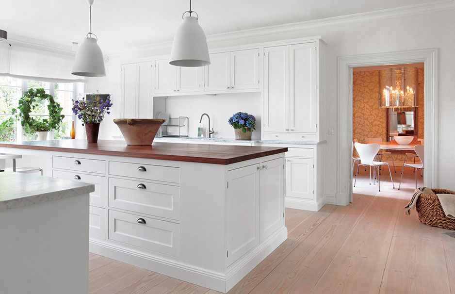 Moderne weiße Landhausküche mit zentraler Kochinsel und Holzakzenten Helle, moderne Landhausküche mit weißer Kücheninsel, Holzarbeitsplatte, Hängelampen und Pflanzen. Im Hintergrund Esszimmer mit Kronleuchter sichtbar.