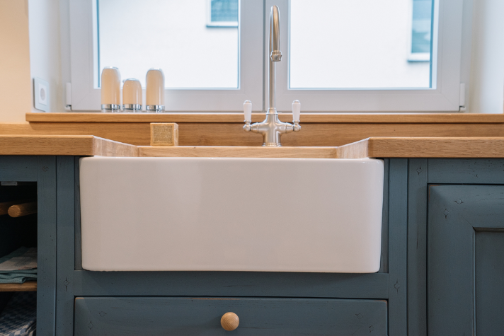 Landhausstil-Küchenspüle aus Keramik mit Wasserhahn und Holzarbeitsplatte Weiße Keramikspüle mit traditionellem Wasserhahn in einer Küche mit Holzarbeitsplatte und blauen Unterschränken