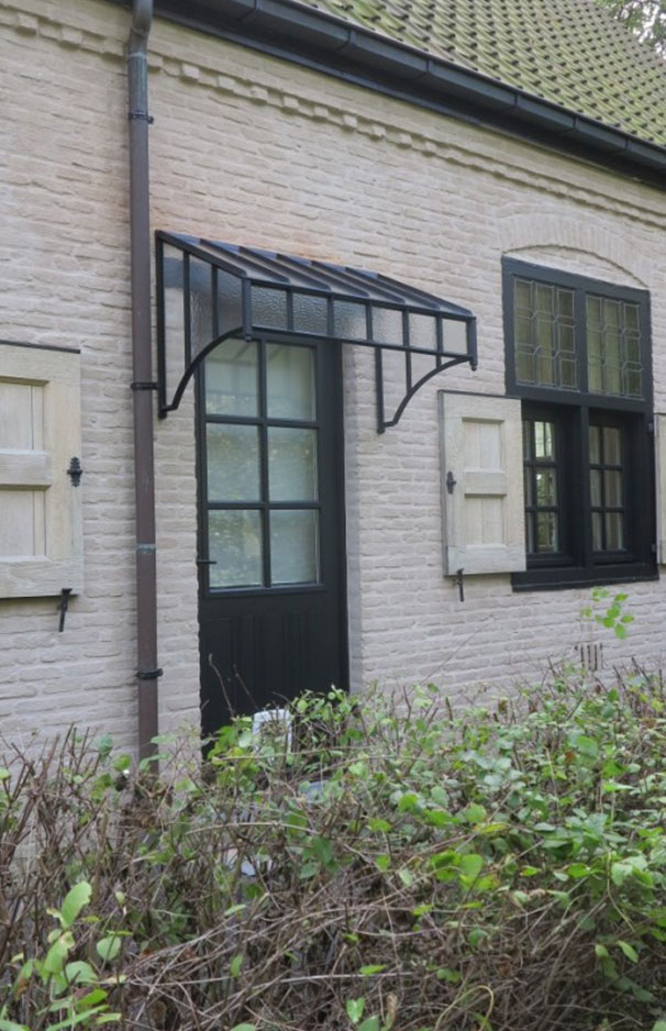 Schwarzes Vordach aus Metall und Glas über einer Haustür an einem Haus mit heller Backsteinfassade Modernes Vordach aus schwarzem Metall und Glas über einer dunklen Haustür in einer Backsteinwand mit weißen Fensterläden