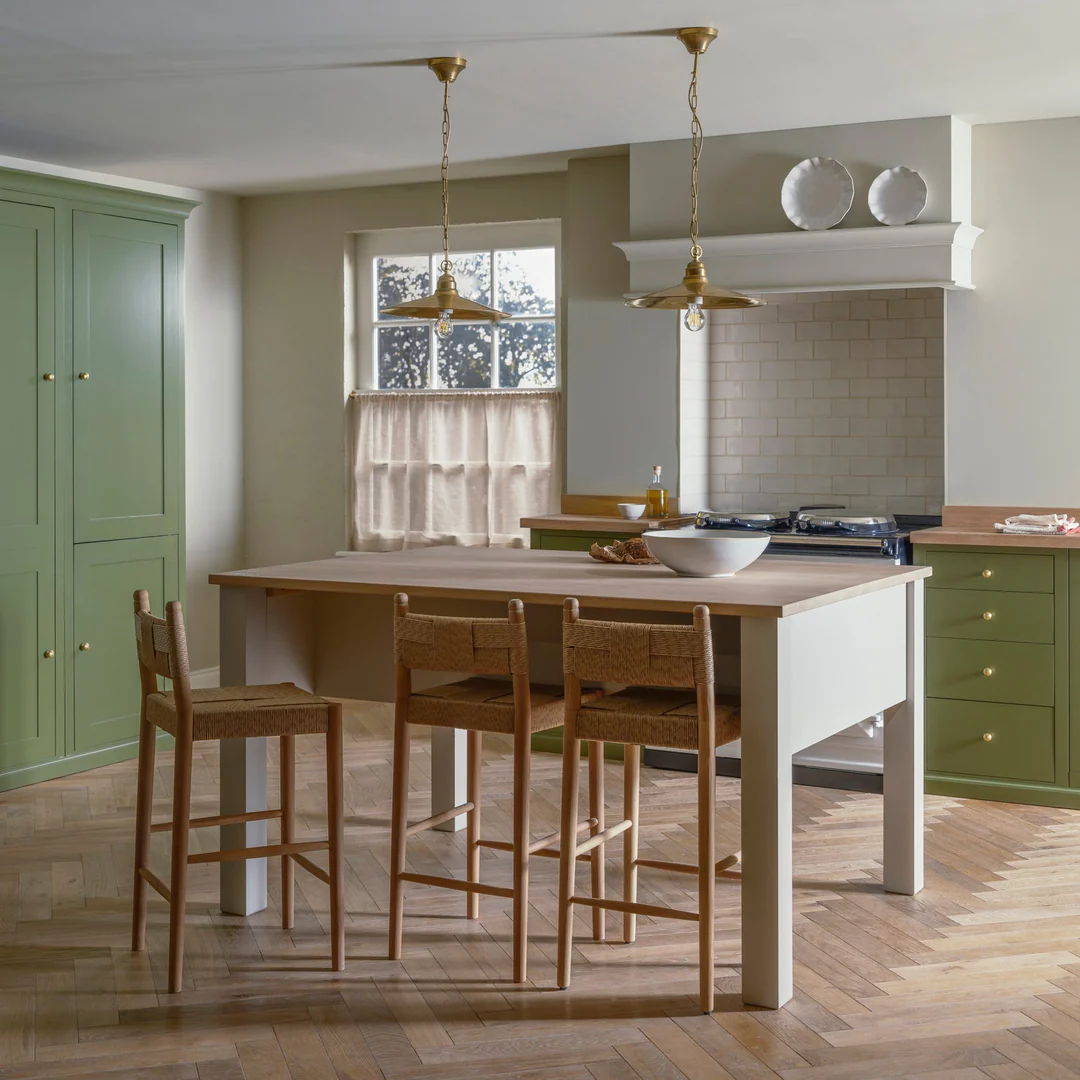 Helle Landhausküche mit salbeigrünen Shaker-Schränken, Messing-Pendelleuchten und großer Kücheninsel aus Holz mit drei Barhockern; Produktfokus: robuste Kücheninsel mit Holzarbeitsplatte.