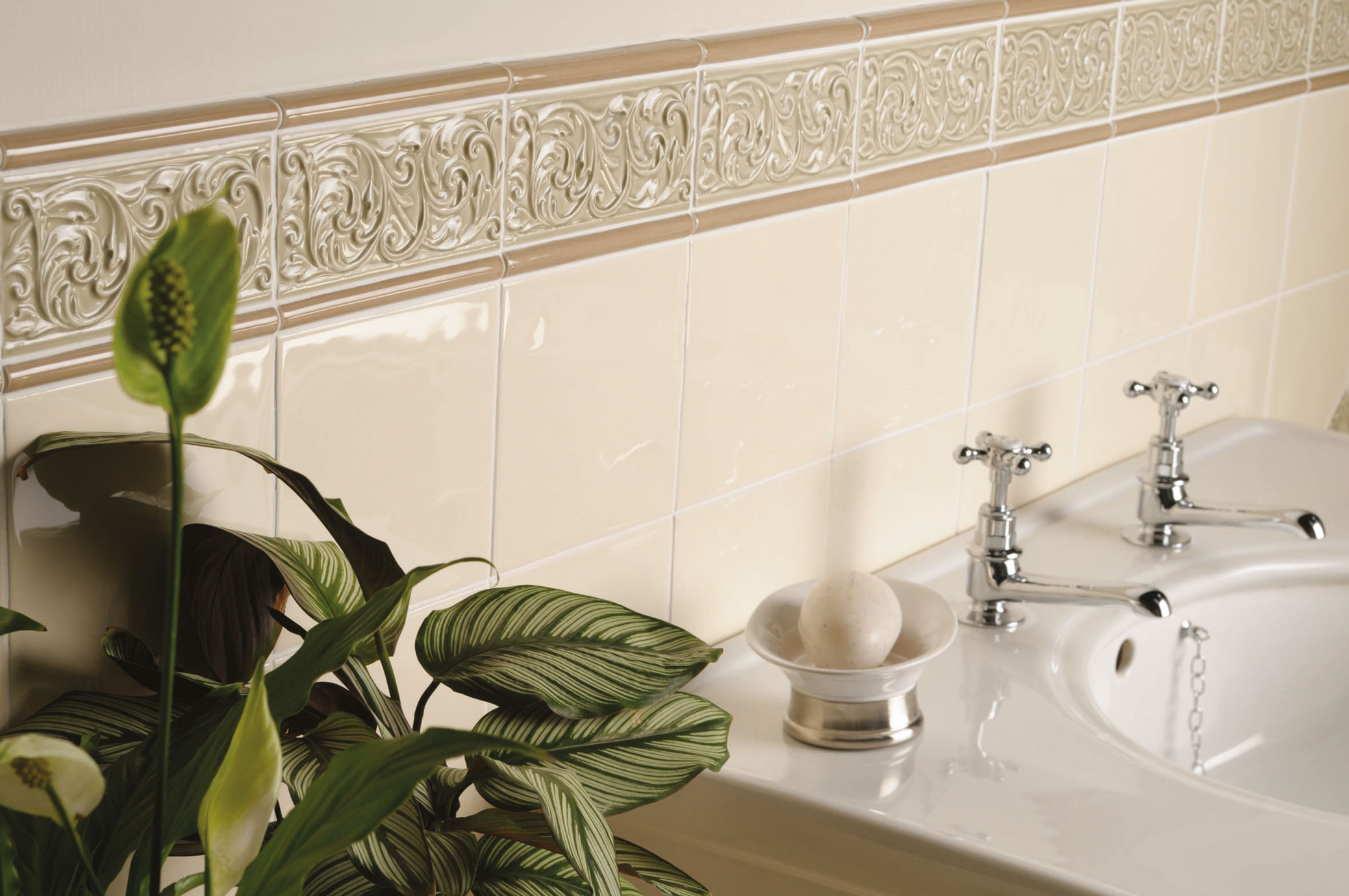 Beige Wandfliesen mit dekorativer Bordüre im klassischen Badezimmer-Design Badezimmer mit glänzenden, beigen Wandfliesen, dekorativer Bordüre, Waschbecken mit zwei Armaturen, Seifenschale und grüner Pflanze