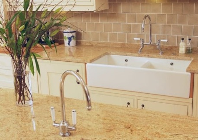 Moderne Landhausküche mit cremefarbener Arbeitsplatte und großer Keramikspüle Große weiße Keramikspüle mit Doppelarmatur in cremefarbener Landhausküche, beige Fliesenrückwand, Blumenstrauß und Küchenutensilien auf der Arbeitsplatte