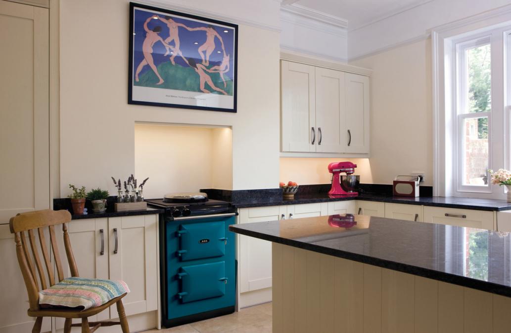Moderne Landhausküche mit cremefarbenen Schränken, schwarzer Granit-Arbeitsplatte, türkisem Herd und pinker Küchenmaschine Helle Landhausküche mit cremefarbenen Schränken, schwarzer Granitplatte, türkisem Herd, Holzstuhl und pinker Küchenmaschine