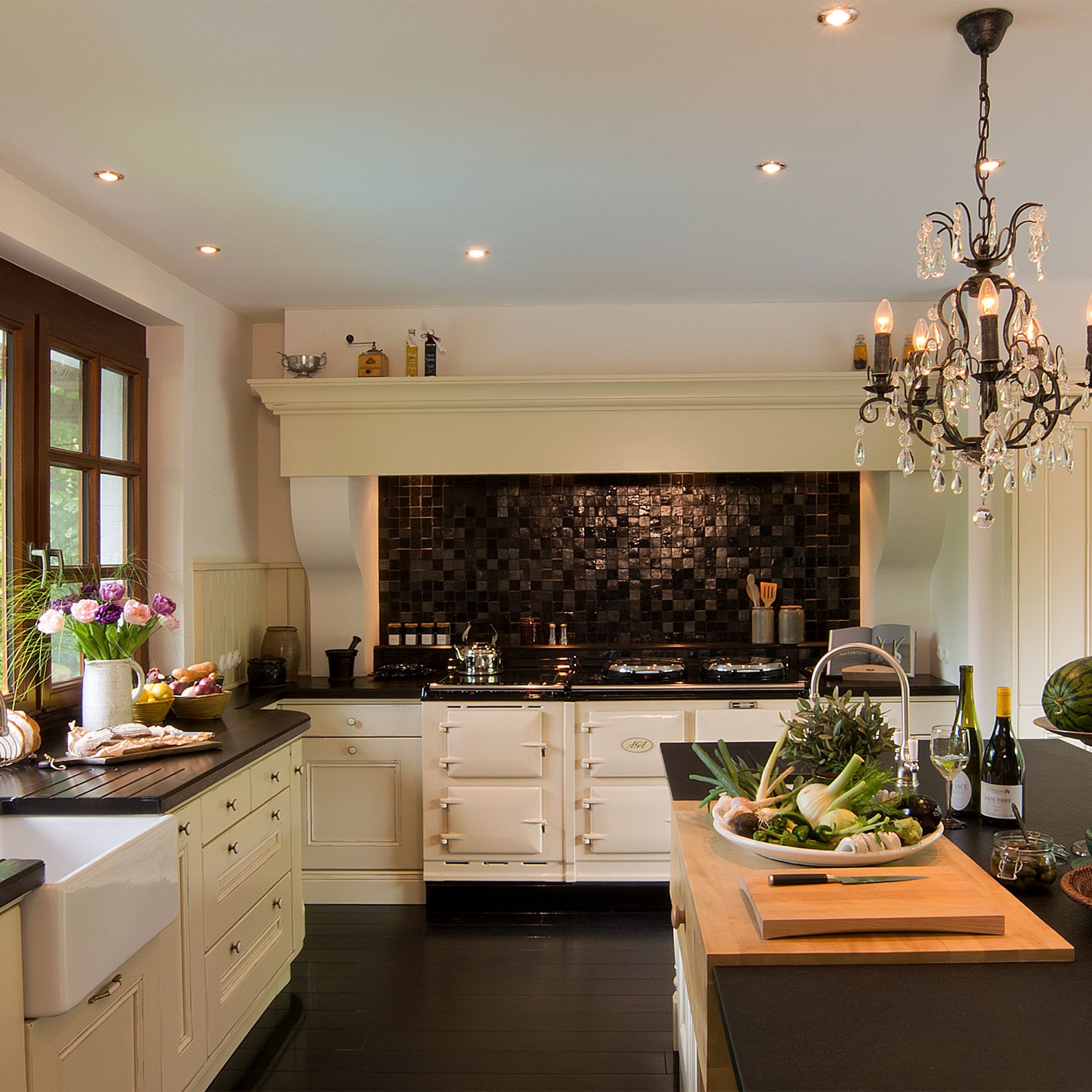 Edle Landhausküche mit Kochinsel, Schneidebrett und Gemüse; cremefarbene Fronten, Spülstein, schwarzer Fliesenspiegel, großer Range Cooker und Kronleuchter – Inspiration für hochwertige Küchenplanung