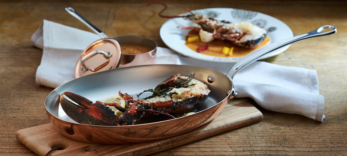 Kupferpfanne mit Deckel, serviert mit frischem Hummer und Zitronenscheiben auf Holzbrett Edle Kupferpfanne mit Deckel, darin ein halbierter Hummer mit Kräutern und Zitronen, im Hintergrund ein angerichteter Teller