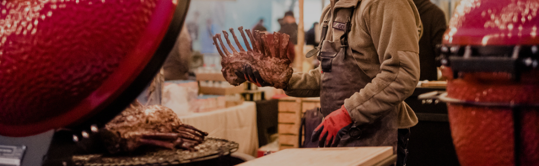 Grillmeister bereitet Lammkarree an einem Keramikgrill auf einem Markt zu Eine Person mit Schürze und roten Handschuhen legt ein Lammkarree auf einen Grill mit rotem Keramikdeckel auf einem Markt