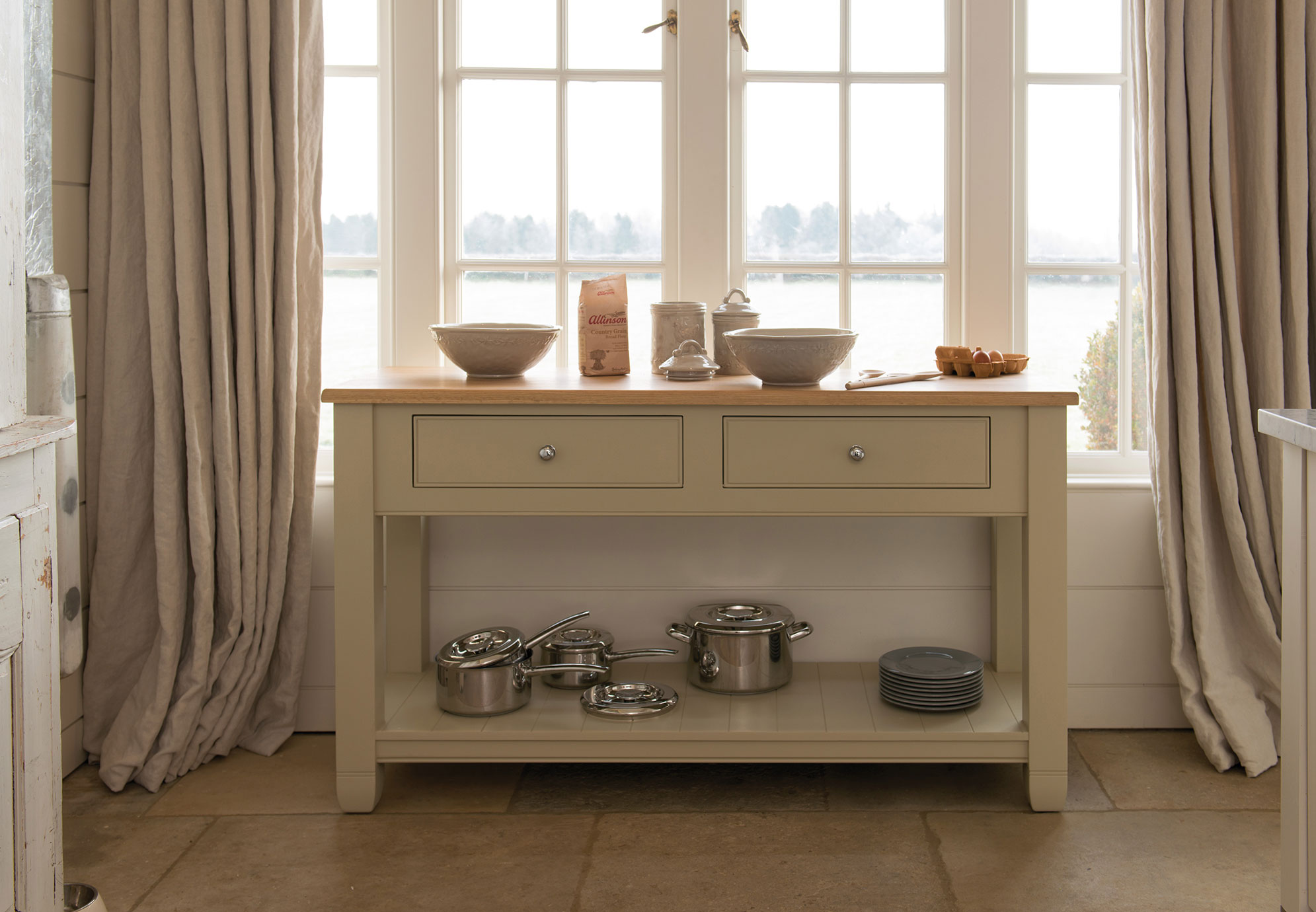 Kücheninsel aus Holz mit Schubladen, Kochgeschirr, Schüsseln und Zutaten vor einem großen Fenster Helle Kücheninsel mit zwei Schubladen, Kochgeschirr und Geschirr auf einer unteren Ablage sowie Schüsseln, Mehl, Eiern und weiteren Küchenutensilien auf der Arbeitsplatte vor einer Fensterfront