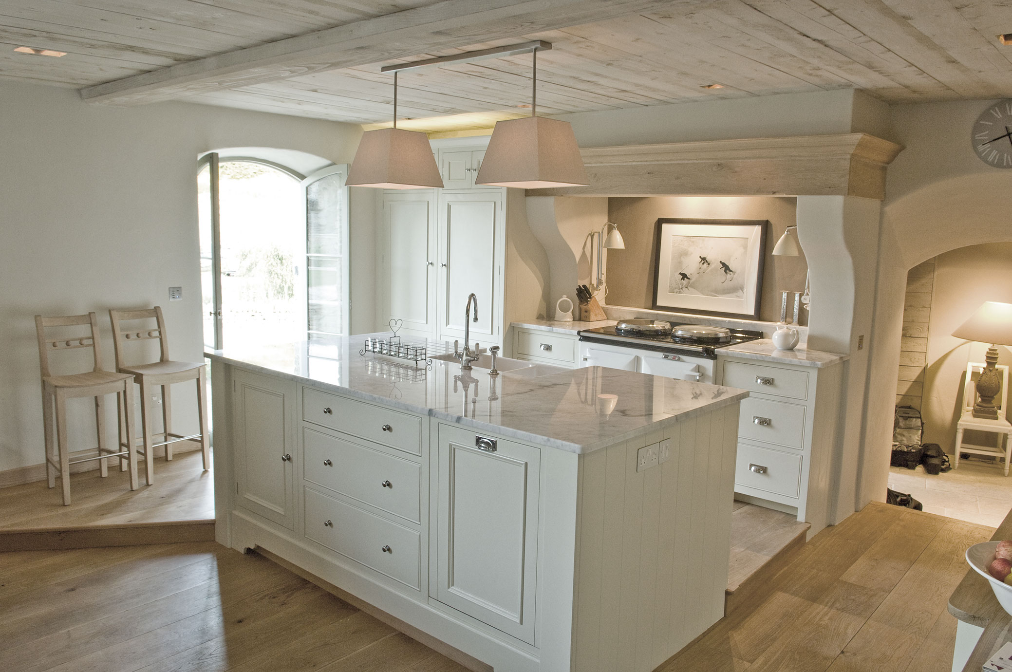 Moderne Landhausküche mit Kochinsel und Marmorarbeitsplatte Helle Landhausküche mit großer Kochinsel, Marmorarbeitsplatte, Hängelampen und zwei Holzstühlen vor einem Fenster