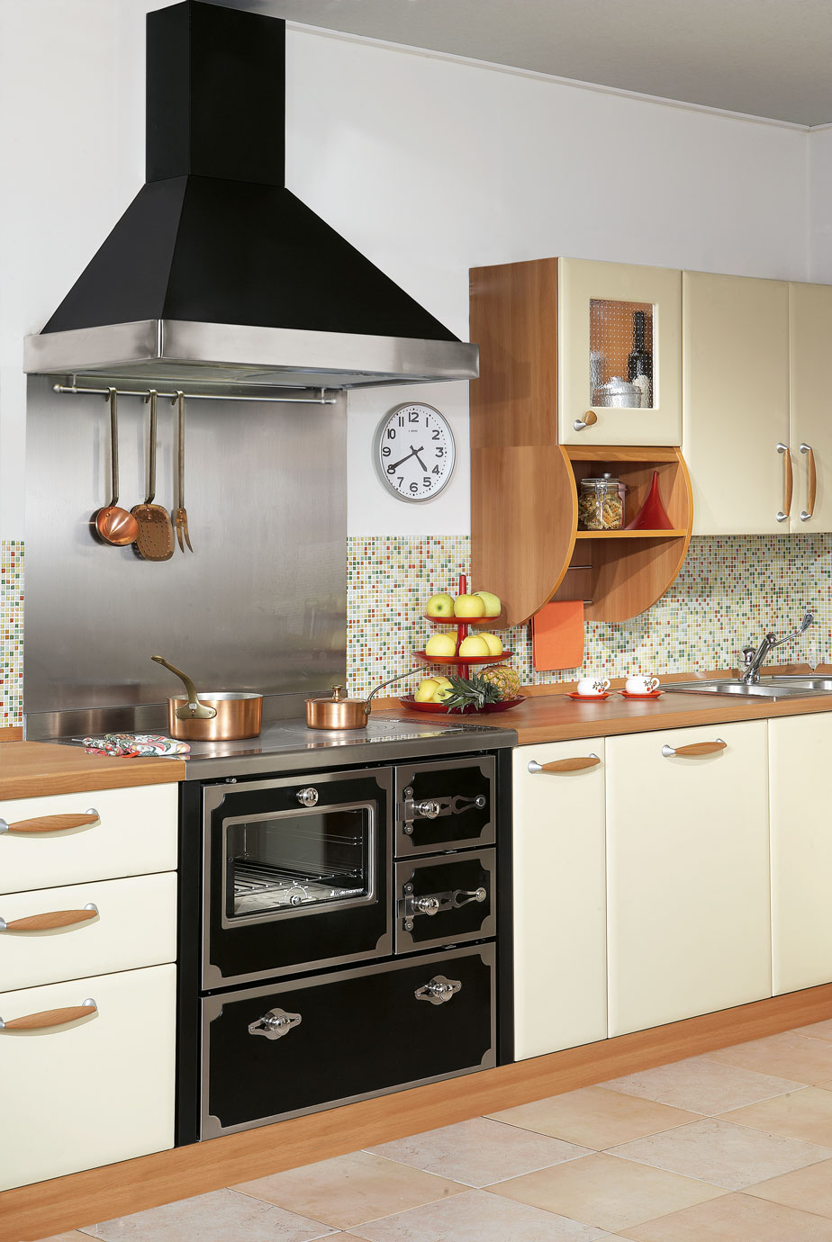 Moderne Einbauküche mit nostalgischem Herd, Kupfertöpfen und bunter Mosaik-Fliese Küchenzeile mit cremefarbenen Schränken, nostalgischem Herd, Dunstabzugshaube, Kupfertöpfen, Obst-Etagere und Wanduhr vor bunter Mosaik-Fliese.