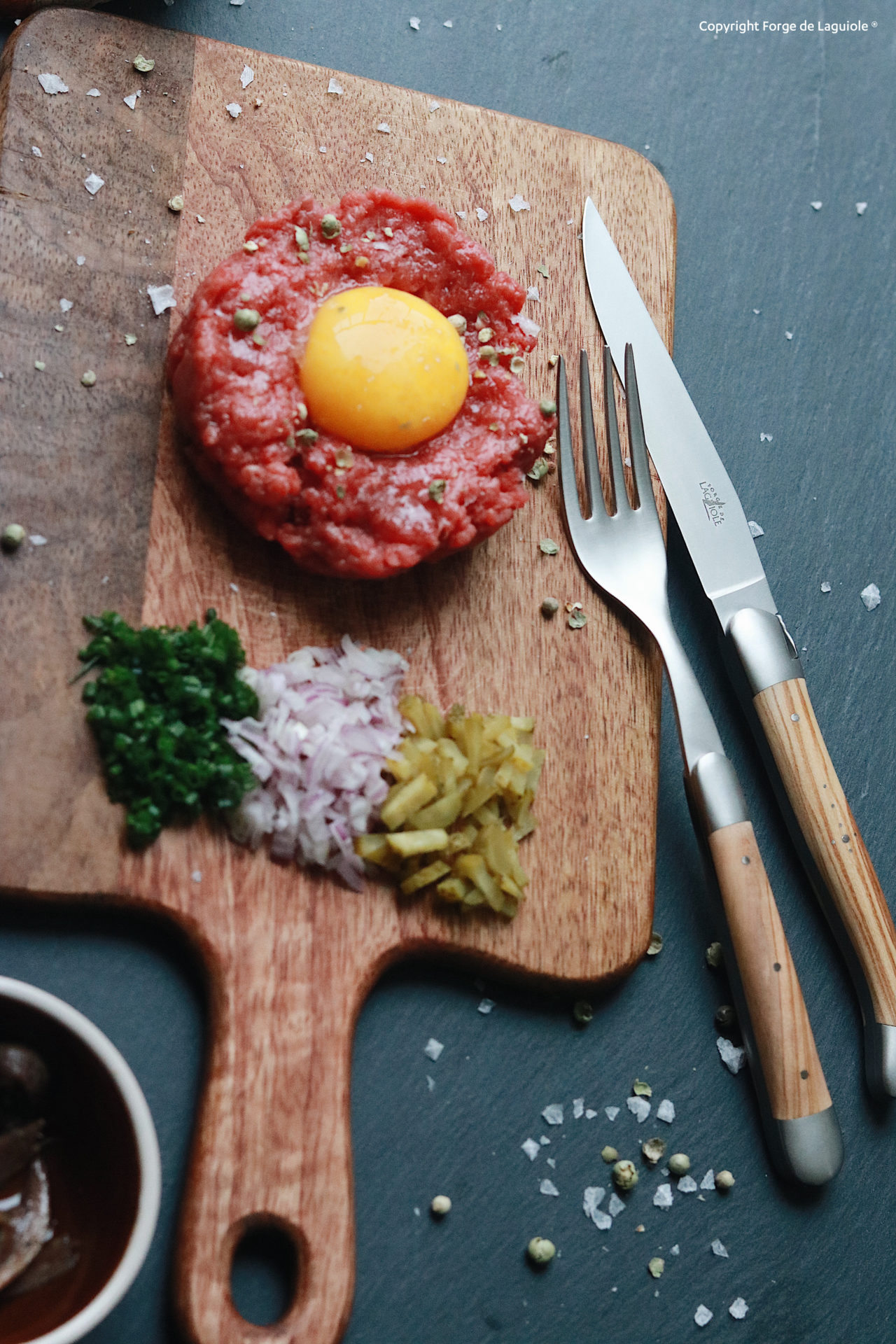 Frisches Beef Tatar mit Eigelb auf Holzbrett, dazu Zwiebeln, Gewürzgurken und Kräuter, angerichtet mit Messer und Gabel Beef Tatar mit rohem Eigelb auf Holzbrett, daneben gehackte Zwiebel, Kräuter und Gewürzgurken, mit Besteck