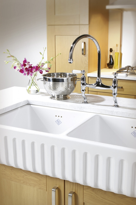 Weiße Doppelspüle aus Keramik mit moderner Armatur in einer stilvollen Küche Doppelspülbecken aus Keramik mit glänzender Chromarmatur, Abtropfsieb und Vase mit lila Orchideen auf einer Arbeitsplatte in einer modernen Küche