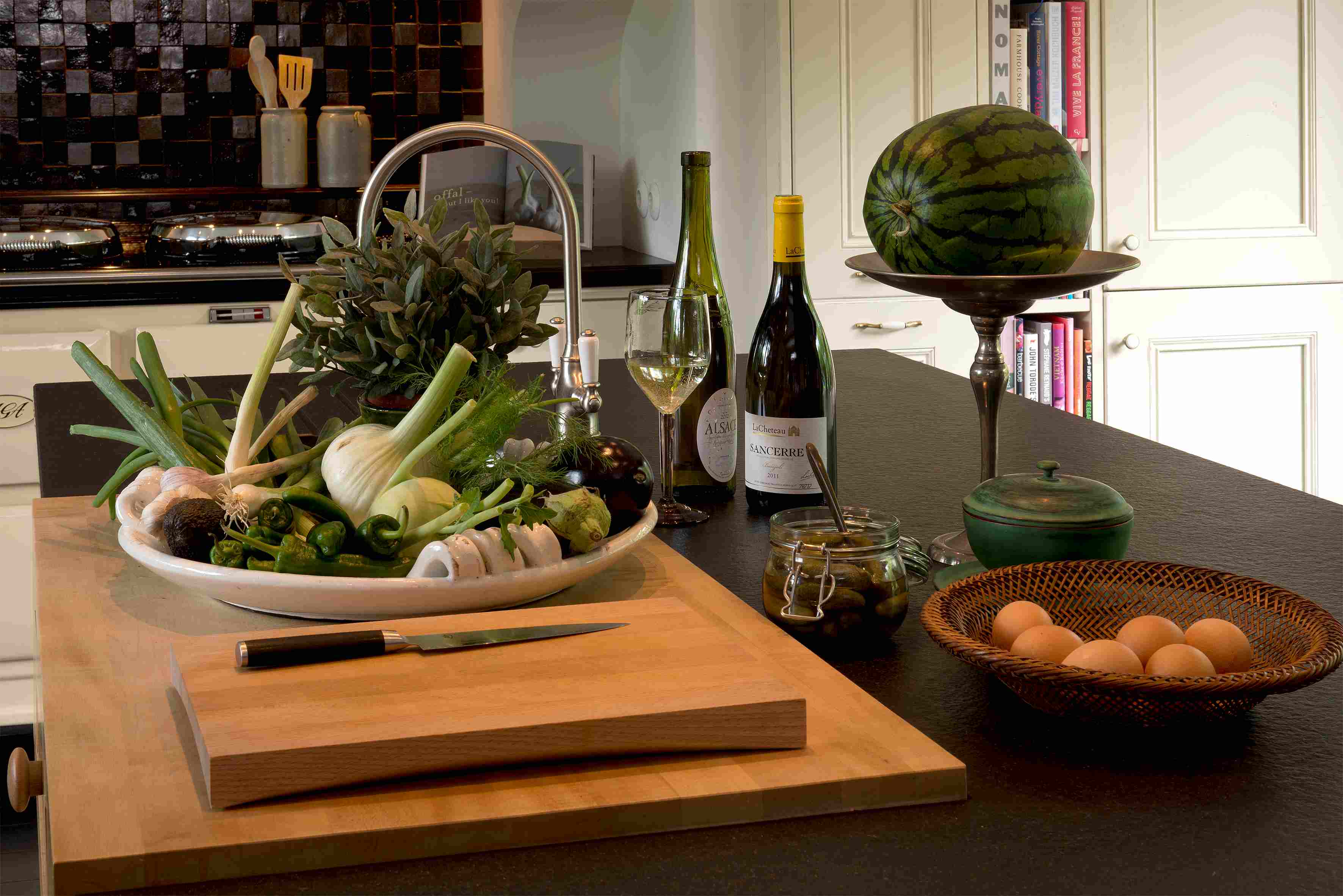 Verschiedene frische Zutaten wie Lauch, Fenchel, Pilze und Kräuter auf einem Schneidebrett, daneben Eier im Korb, Einmachglas, zwei Flaschen Wein, Weinglas und eine große Wassermelone in einer modernen Küche