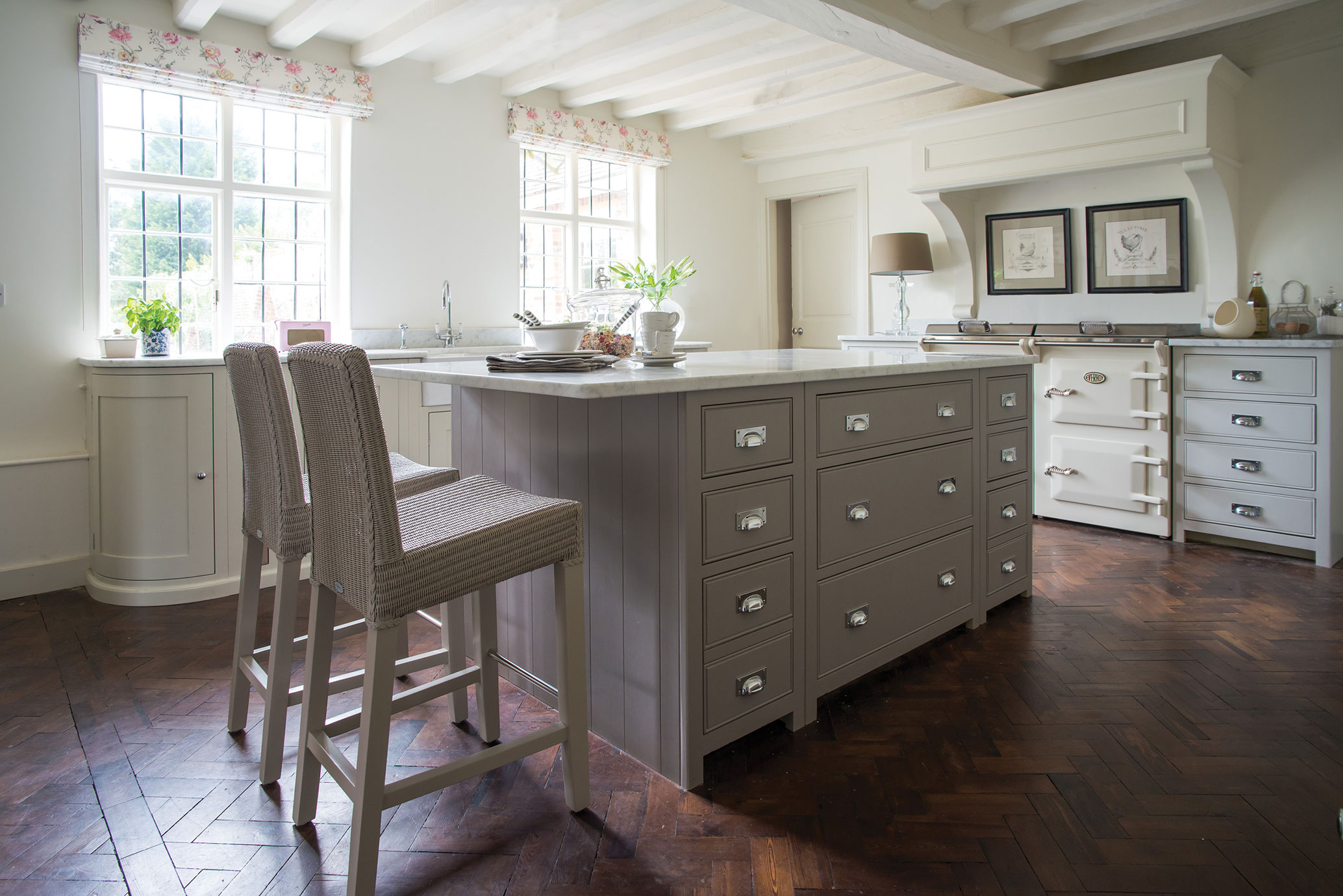 Landhausstil-Küche mit Kücheninsel, Barhockern und großen Fenstern – helle, einladende Atmosphäre Helle Landhausküche mit grauer Kücheninsel aus Holz, zwei geflochtenen Barhockern, weißen Schränken, großem Herd, Parkettboden und großen Fenstern