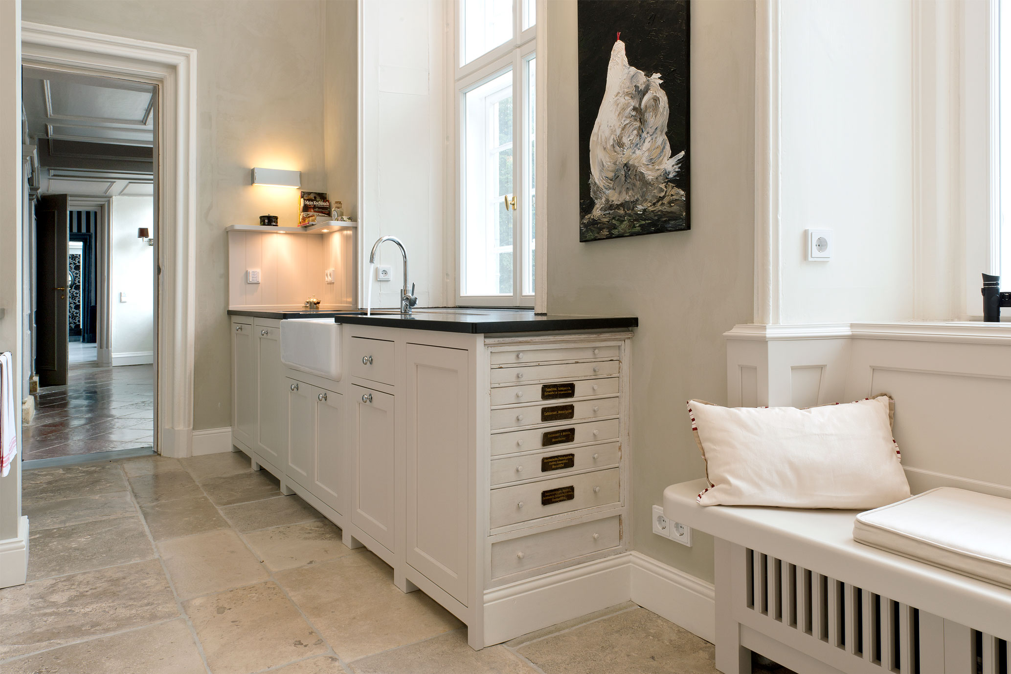 Helle, moderne Landhausküche mit weißen Schränken, Spülbecken und gemütlicher Sitzbank Elegante Küche im Landhausstil mit weißen Fronten, schwarzen Arbeitsflächen, Spülbecken, Sitzbank mit Kissen und Huhn-Gemälde an der Wand