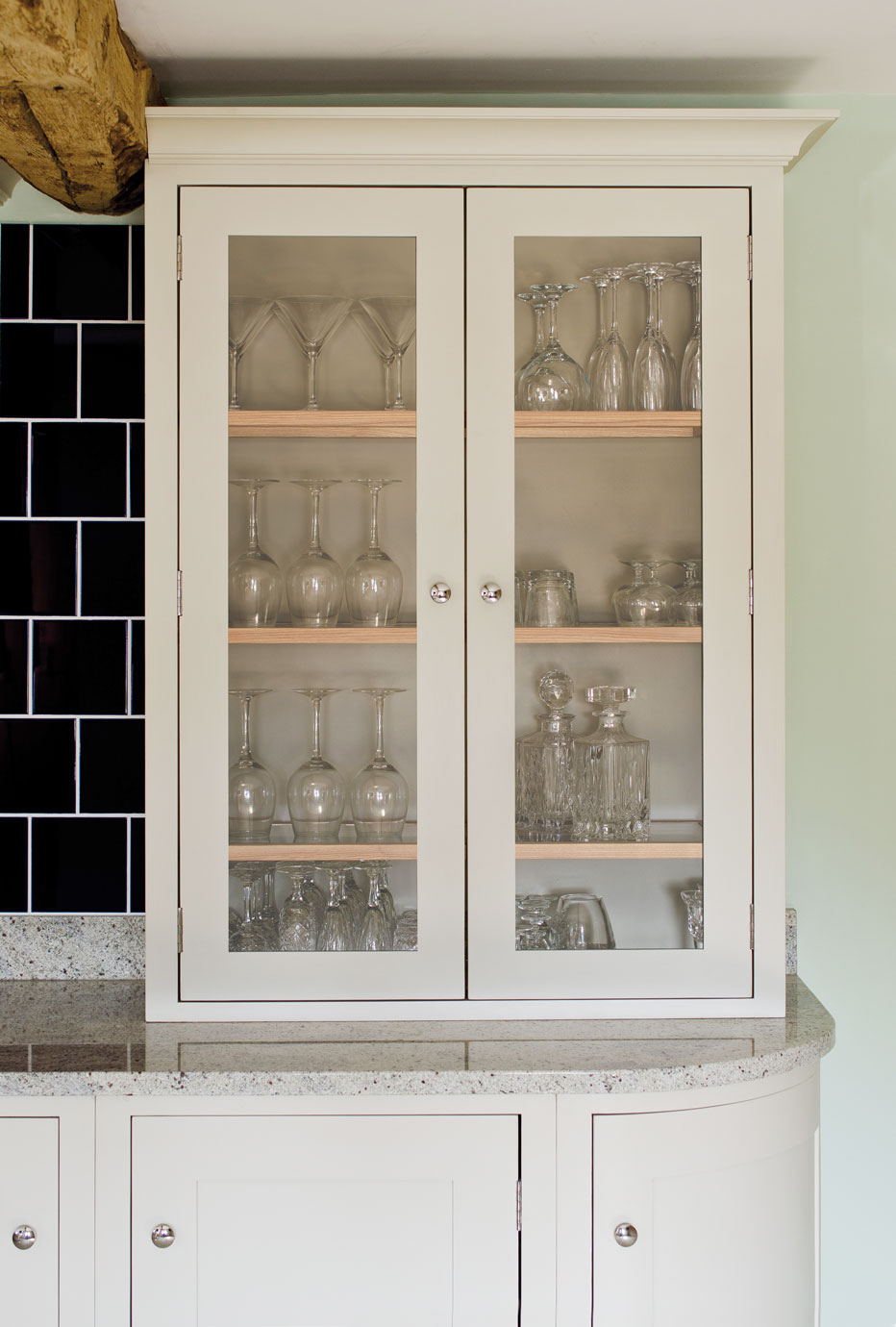 Eleganter Vitrinenschrank aus Holz mit Glastüren und Gläsern, ideal für moderne Küchen Heller Vitrinenschrank aus Holz mit Glastüren, gefüllt mit verschiedenen Gläsern und Karaffen, auf einer Granit-Arbeitsfläche stehend