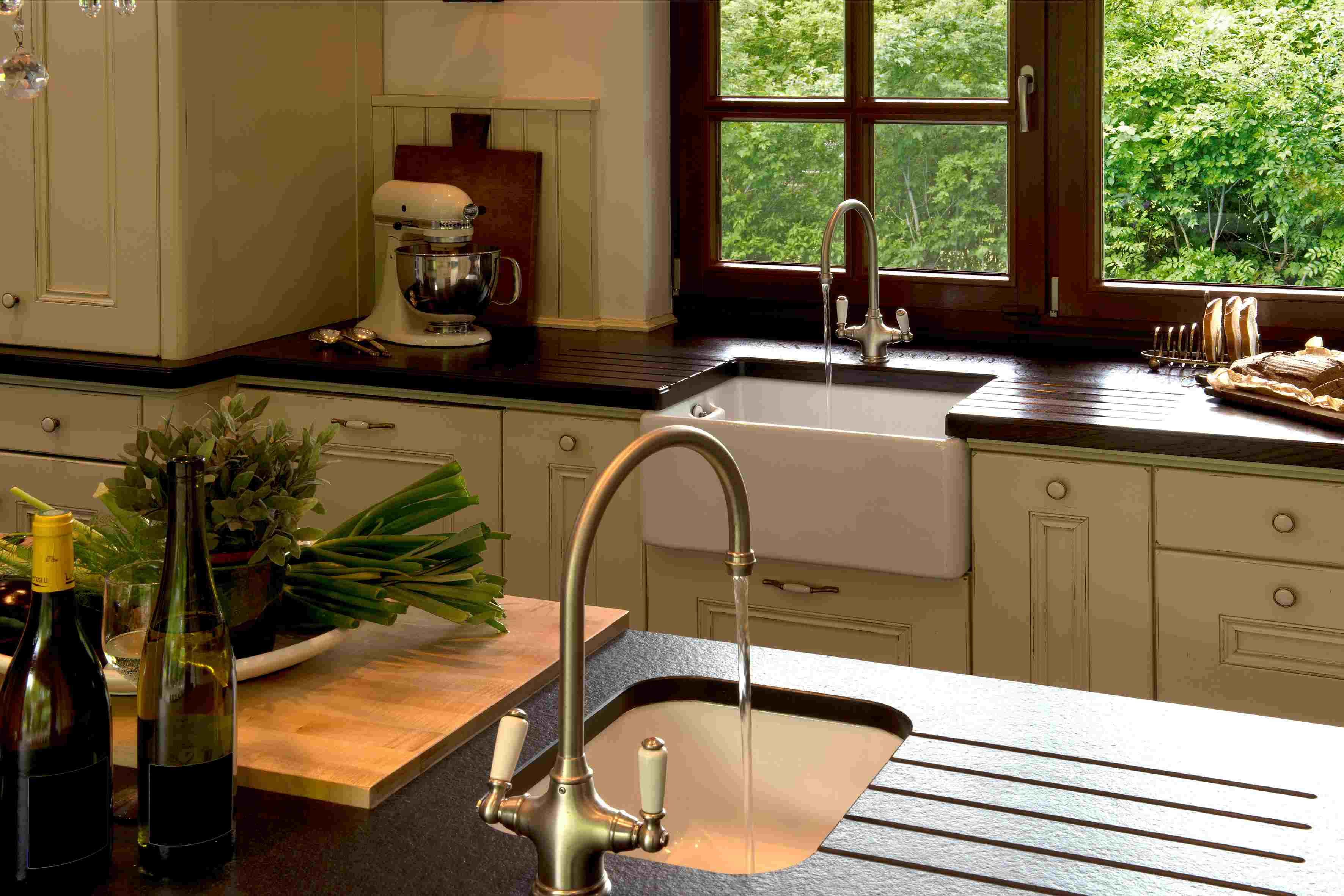 Gemütliche Küche im Landhausstil mit zwei Spülen, Küchenmaschine, Holzarbeitsplatte, frischen Kräutern, Weinflaschen und großem Fenster mit Blick auf Garten