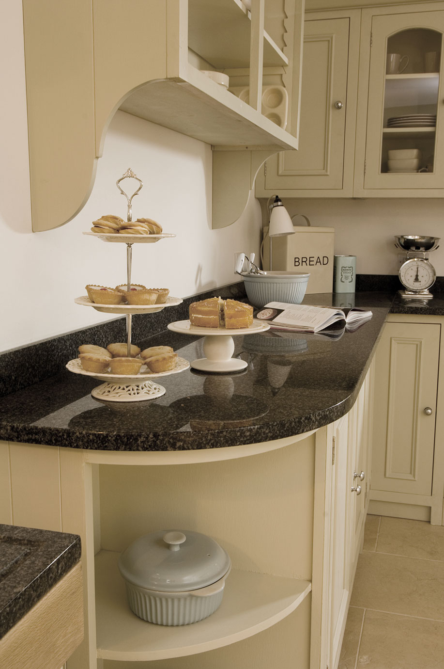 Elegante Landhausküche mit Granit-Arbeitsplatte und frischem Gebäck auf Etagere Helle Küche im Landhausstil mit Granitarbeitsplatte, Etagere mit Gebäck und Kuchen, Küchenwaage, Brotkasten und Kochbuch