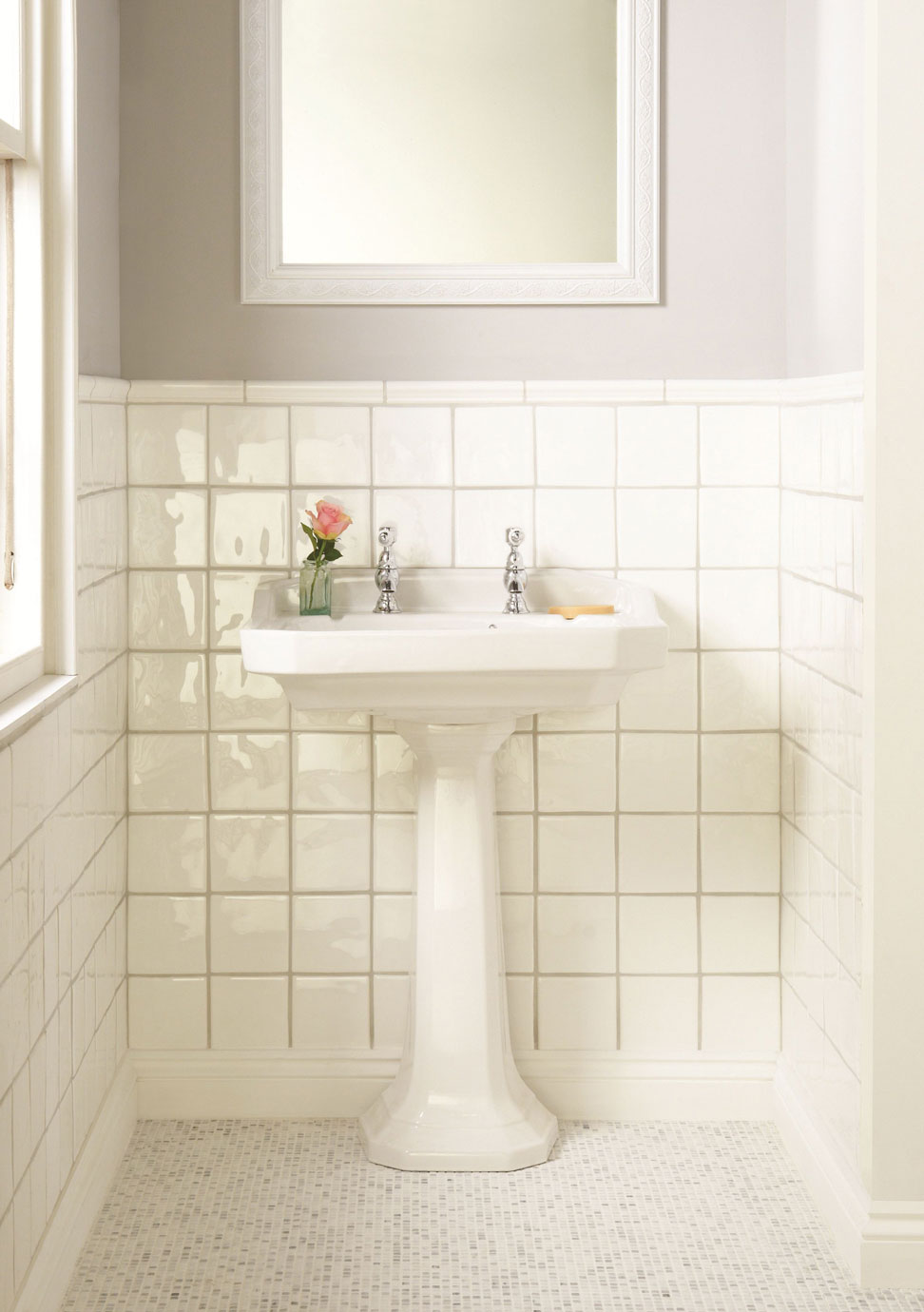 Weißes Standwaschbecken mit zwei Armaturen, einer Seifenschale und einer rosa Blume in einer Vase vor hellen Wandfliesen im Bad