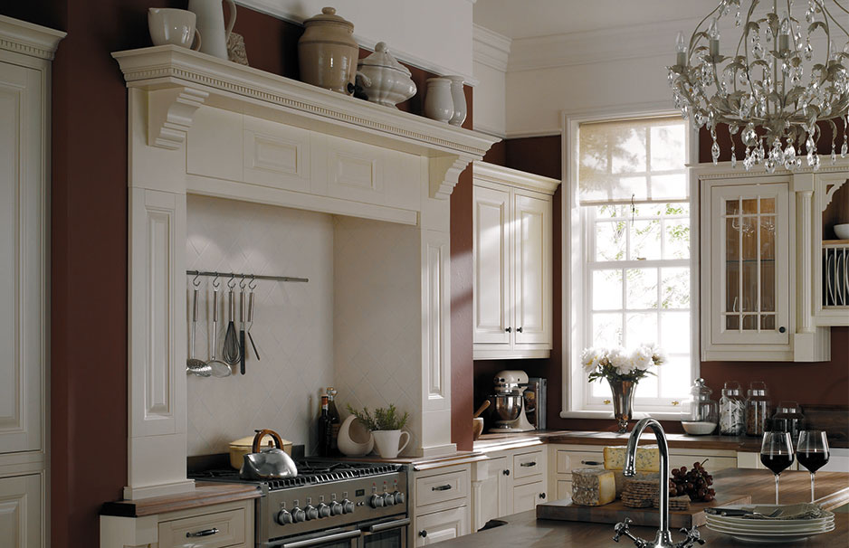 Klassische Landhausküche mit weißen Schränken, Gaskochfeld, Kronleuchter und großen Fenstern Elegante Landhausküche mit cremefarbenen Küchenmöbeln, Kochbereich, Kronleuchter und Fenster, dekoriert mit Blumen und Küchenutensilien