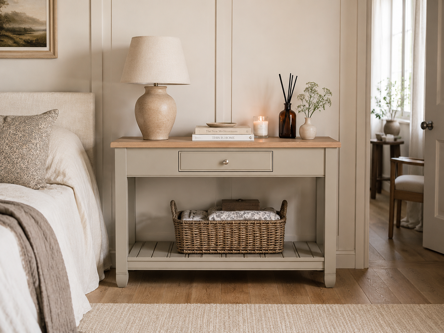 Beiger Landhaus-Konsolentisch aus Holz mit Eichenplatte, Schublade und unterem Regal, dekoriert mit Lampe, Büchern, Duftstäbchen und Kerze; Korb auf der Ablage – ideal als Sideboard im Schlafzimmer.