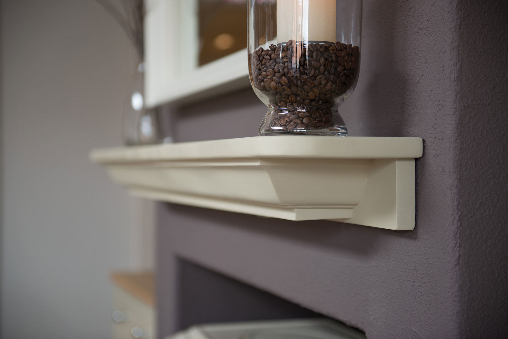 Elegante cremefarbene Kaminkonsole aus Holz an einer lila Wand mit Glasvase, gefüllt mit Kaffeebohnen und Kerze als Dekoration Detailaufnahme einer cremefarbenen Kaminkonsole aus Holz, dekoriert mit einer Glasvase voller Kaffeebohnen und einer Kerze an einer lila Wand