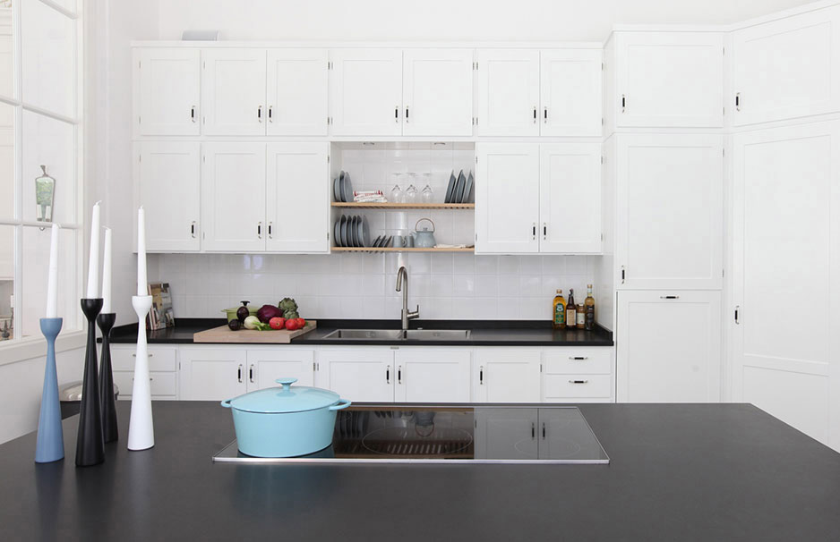 Moderne weiße Küche mit schwarzer Arbeitsplatte, Kochinsel und Dekoelementen Helle, moderne Küche mit weißen Schränken, schwarzer Arbeitsfläche, Kochfeld, blauer Kasserolle, Kerzenständern und frischem Gemüse auf der Arbeitsplatte