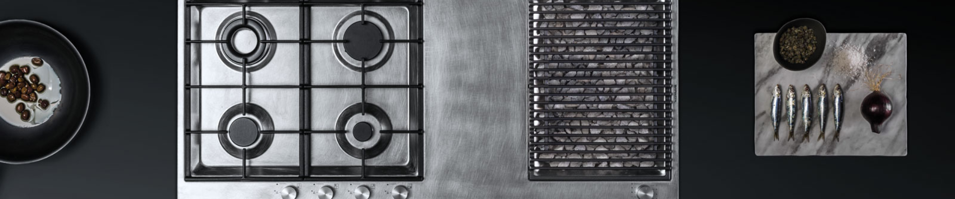 Edelstahl-Gaskochfeld mit vier Brennern und Grillfunktion, daneben Zutaten auf Marmorbrett arrangiert Modernes Gaskochfeld aus Edelstahl mit Grillfläche, daneben Oliven in einer Pfanne sowie Sardinen, Zwiebel und Gewürze auf einem Marmorbrett