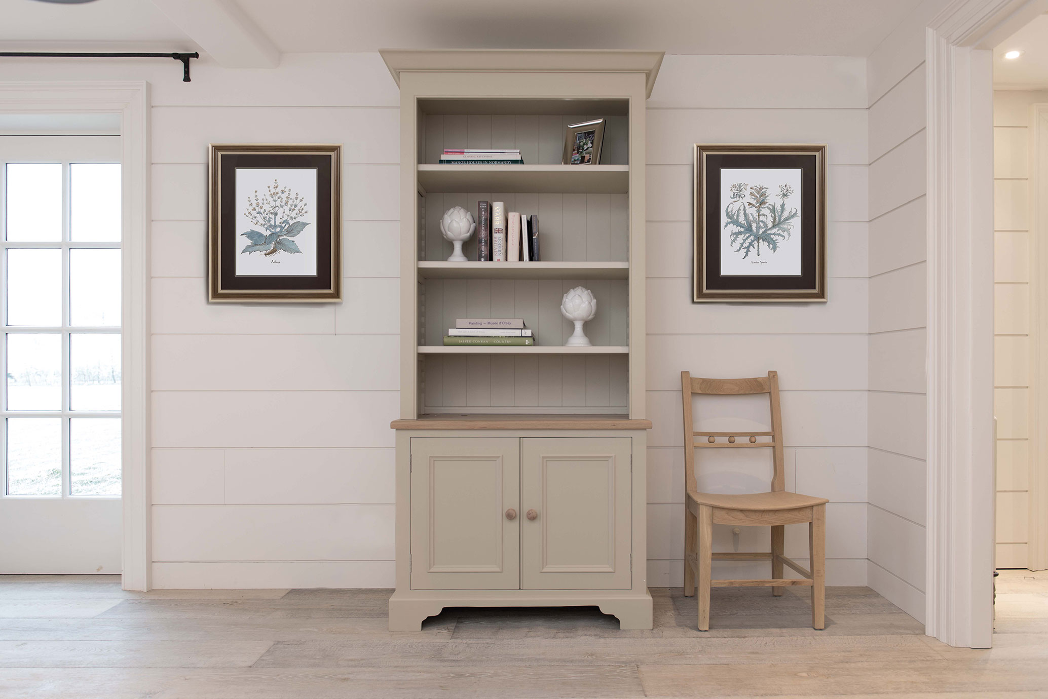 Landhausstil Regal mit Schrank und Bücherregal in hellem Wohnzimmer mit dekorativen Bildern und Holzstuhl Beigefarbenes Regal mit offenen Fächern und Schranktüren in einem hellen Raum, dekoriert mit Büchern, weißen Deko-Objekten, gerahmten botanischen Bildern und einem Holzstuhl