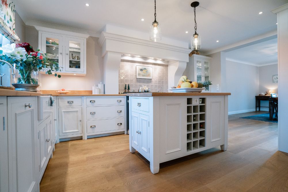 Elegante weiße Landhausküche: Kücheninsel mit Weinregal, Holz-Arbeitsplatten, Hängeschränke mit Glas und Pendelleuchten – ideale Inspiration für hochwertige Einbauküchen
