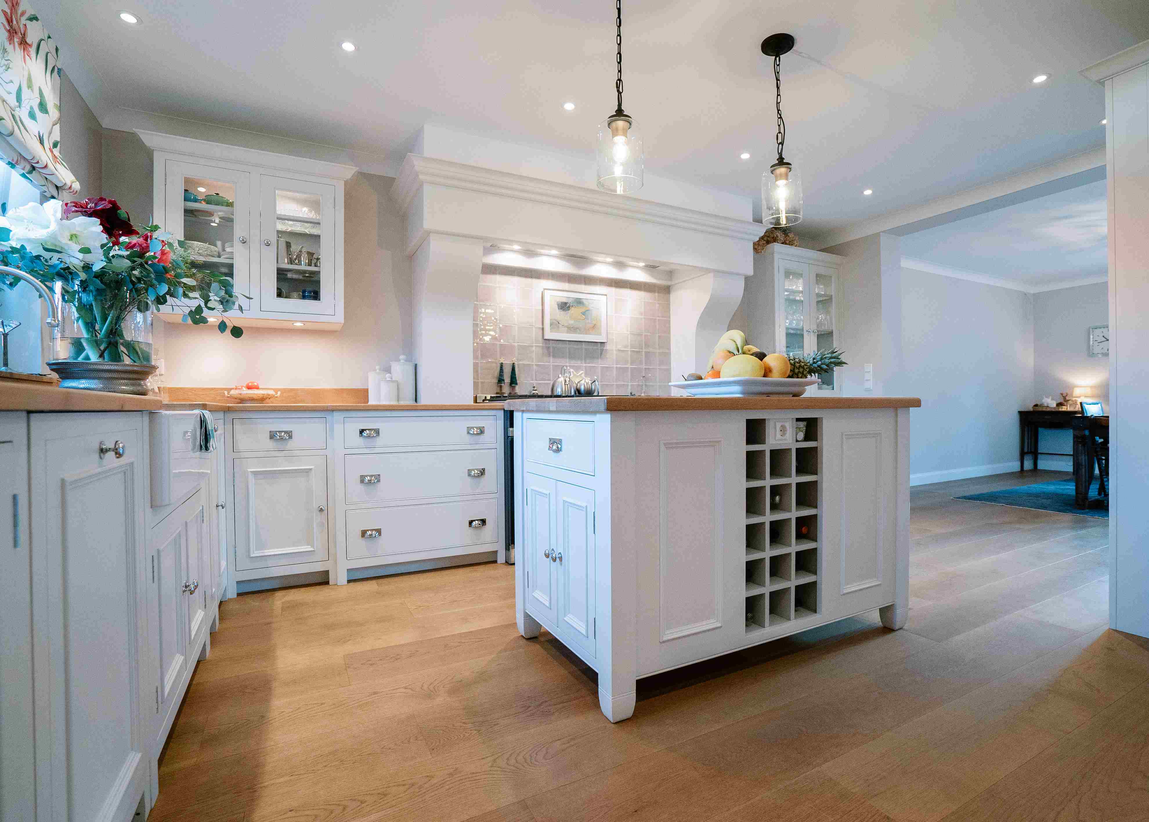 Helle, großzügige Küche im Landhausstil mit Kücheninsel, weißen Holzfronten, Obstschale und eleganter Arbeitsfläche auf hellem Parkettboden