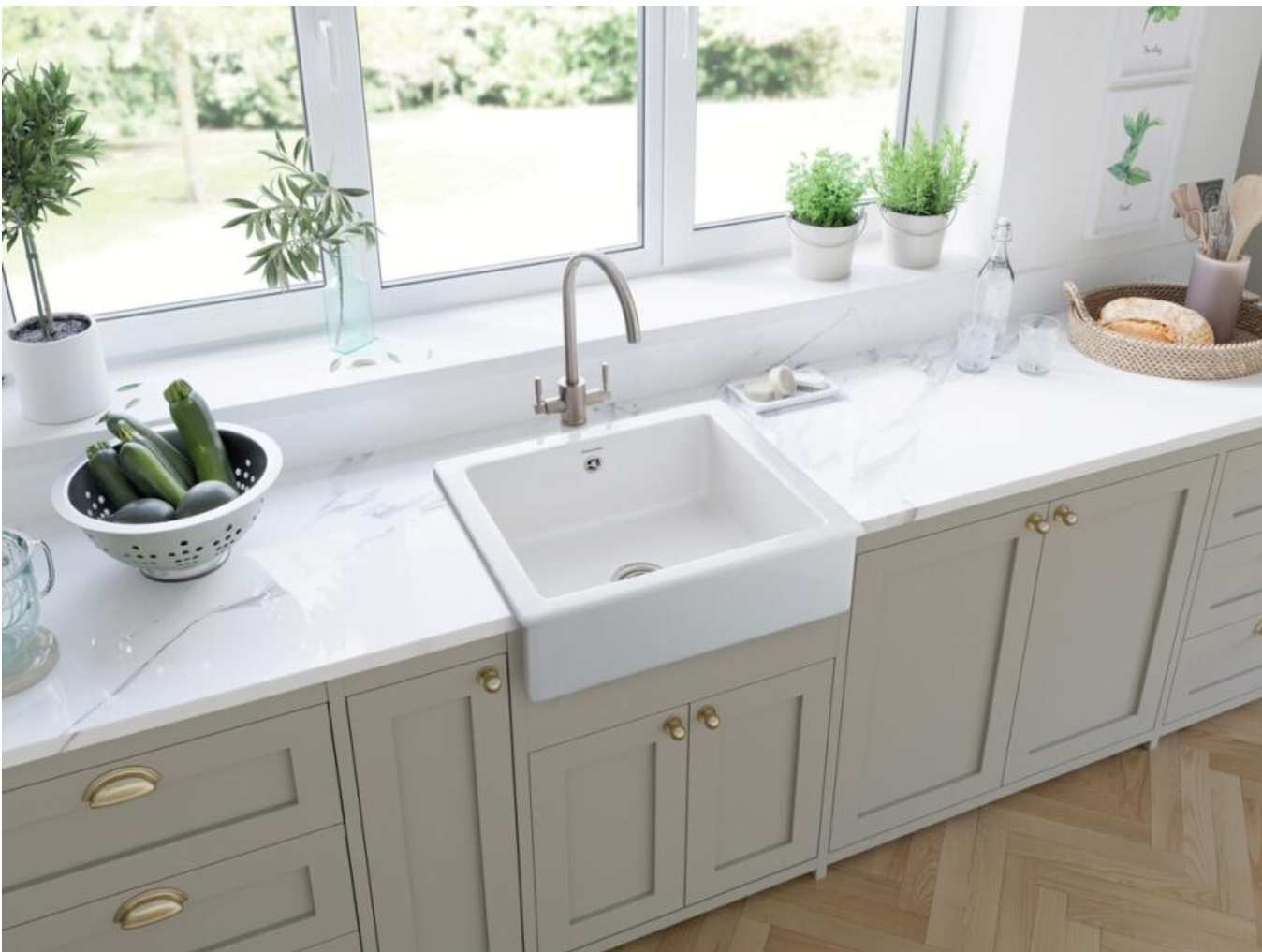 Weiße apron-front Keramik-Küchenspüle mit hoher Armatur in heller Küche mit Arbeitsplatte in Marmor-Optik, Kräutertöpfen auf der Fensterbank und Sieb mit Zucchini