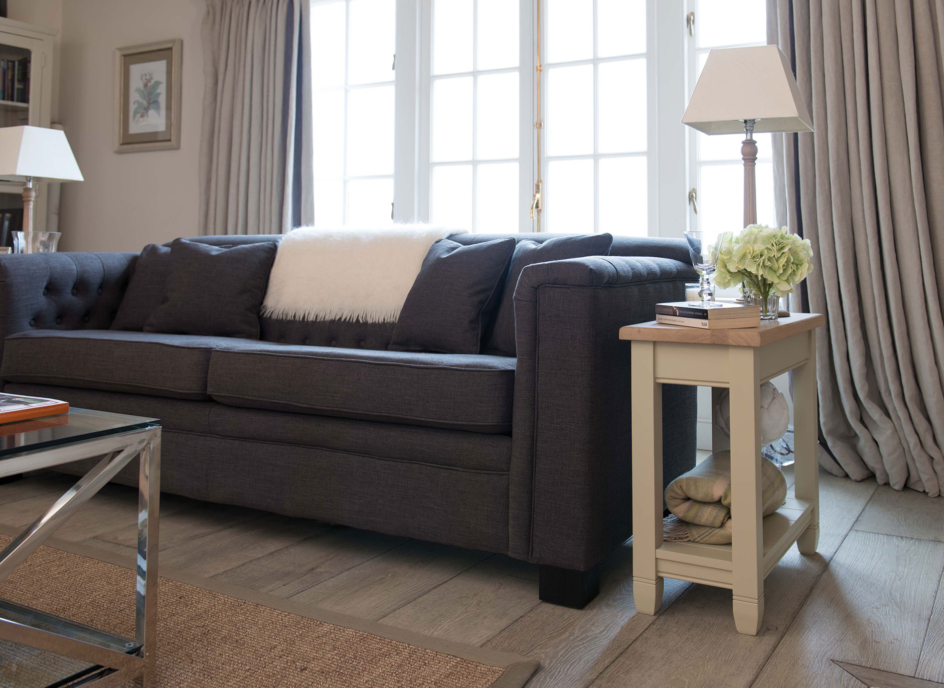 Modernes Wohnzimmer mit dunkelgrauem Sofa und stilvollem Beistelltisch Gemütliches Wohnzimmer mit dunkelgrauem Sofa, Kissen, weißem Fell und Beistelltisch mit Lampe und Blumen