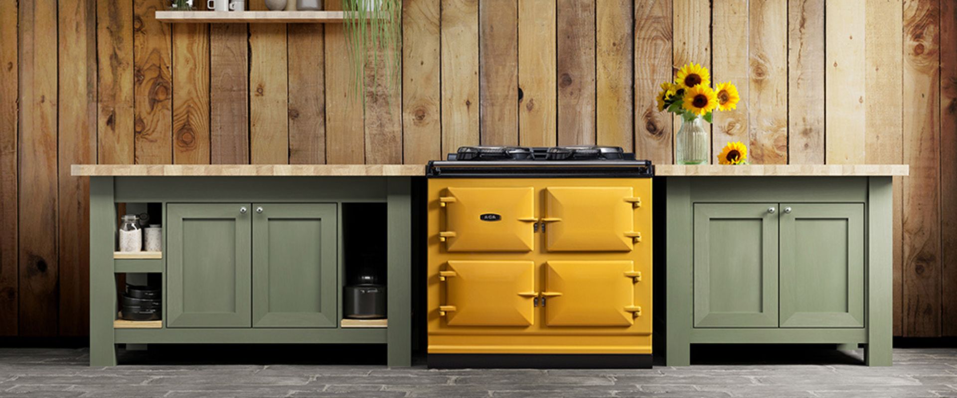 Gelber Range Cooker mit vier Ofentüren und Gaskochfeld, eingebaut zwischen salbeigrünen Unterschränken mit Holzarbeitsplatte; Deko mit Sonnenblumen – stilvoller Blickfang für die Landhausküche.