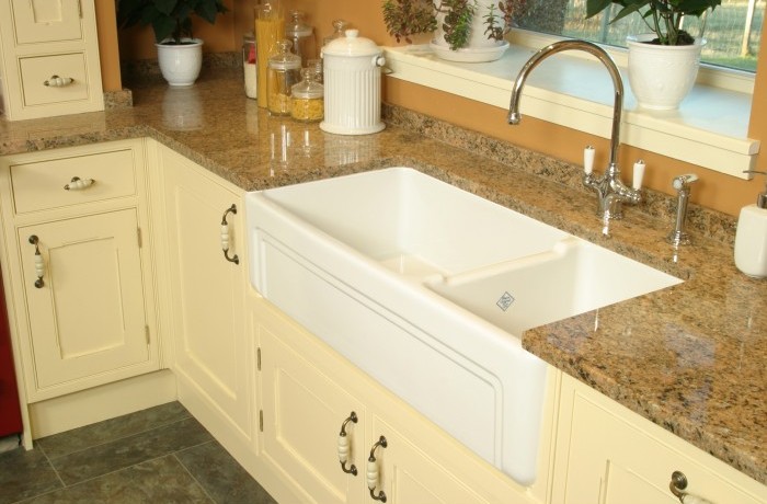 Moderne Landhausküche mit Keramikspüle und Granit-Arbeitsplatte Geräumige weiße Keramikspüle in einer cremefarbenen Landhausküche mit Granit-Arbeitsplatte, Wasserhahn und dekorativen Utensilien auf der Fensterbank.