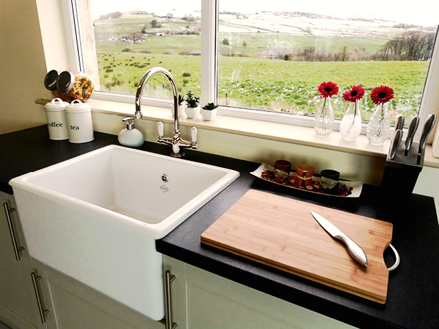 Moderne Küchenzeile mit Spülbecken, Schneidebrett und Blick ins Grüne Helle Küche mit großem Spülbecken, Holzschneidebrett, Messer, Kaffeedosen und Blick auf eine grüne Landschaft durch das Fenster