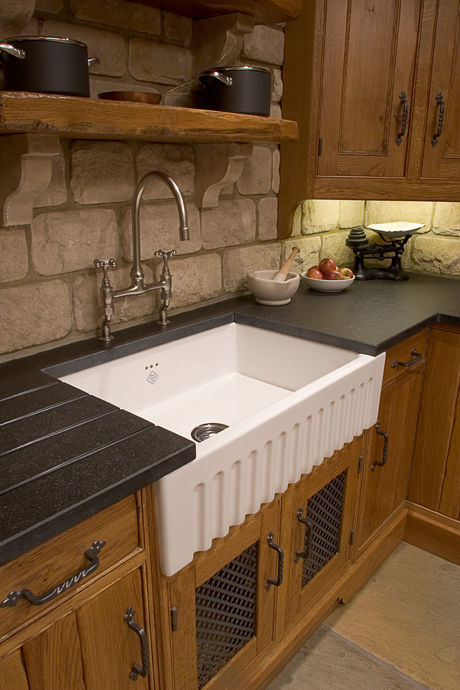 Landhausstil-Küchenspüle aus Keramik mit traditioneller Armatur und Holzunterschrank Weiße Keramikspüle im Landhausstil mit moderner Armatur, umgeben von Natursteinwand und Holzschränken in einer gemütlichen Küche