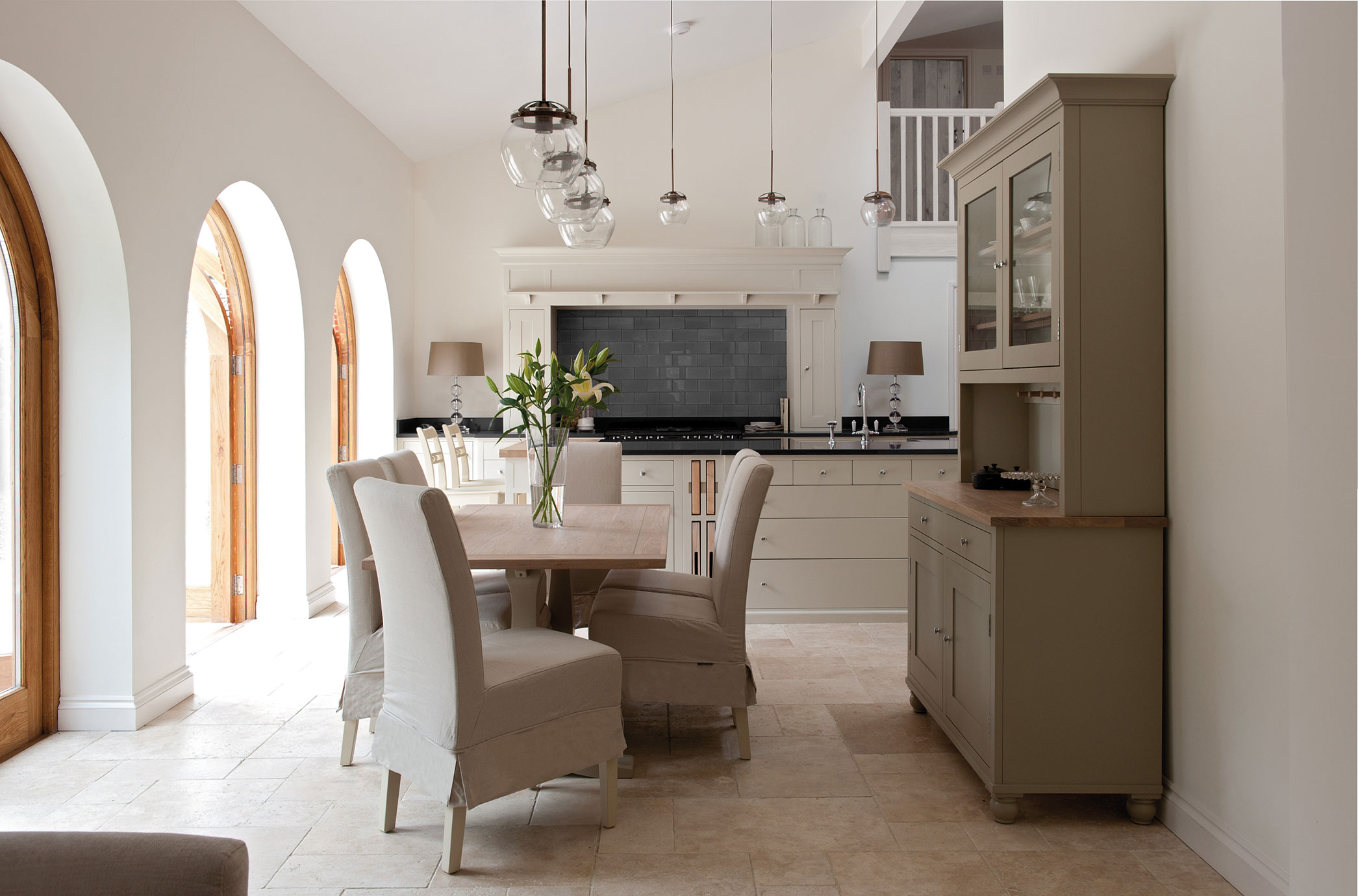 Moderne Landhausküche mit großen Fenstern und Essbereich Helle Landhausküche mit Esstisch, gepolsterten Stühlen, Vitrinenschrank, Hängelampen und großen Rundbogenfenstern aus Holz