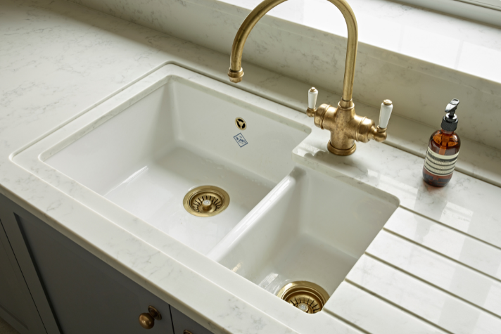 Doppelspülbecken aus Keramik mit goldener Armatur in moderner Küche Elegantes weißes Keramik-Doppelspülbecken mit goldfarbenen Abflüssen und passender Küchenarmatur, daneben eine Seifenspenderflasche auf heller Arbeitsplatte