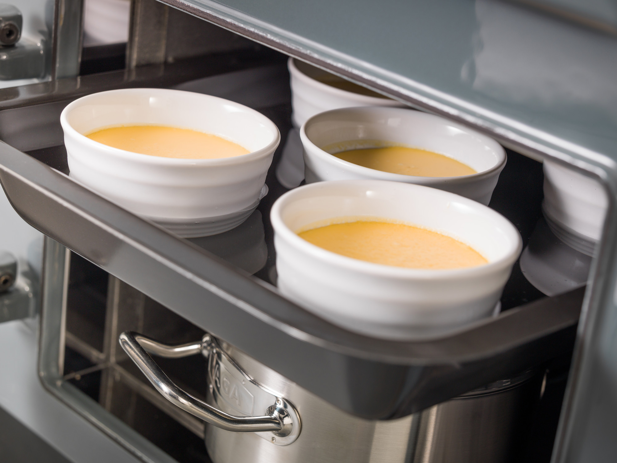 Weiße Keramikförmchen mit Creme-Dessert im Ofen auf Backblech Mehrere weiße Keramikförmchen gefüllt mit gelber Creme stehen auf einem Backblech im geöffneten Ofen.