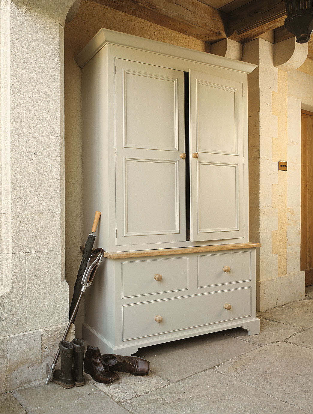 Großer, cremefarbener Garderobenschrank mit Holzakzenten und Schubladen im Flur Eleganter Garderobenschrank aus Holz mit zwei Türen, zwei großen Schubladen und Holzgriffen. Davor stehen Regenschirm, Reitstiefel und Schuhe auf Steinboden.