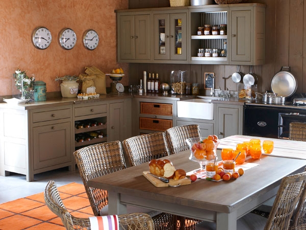 Gemütliche Landhausküche mit Holzmöbeln, Esstisch und frischen Backwaren Einladende Landhausküche mit gedecktem Holztisch, Rattanstühlen, Glasgefäßen mit Marmelade und frischem Brot auf dem Tisch, im Hintergrund Küchenmöbel und Wanduhren
