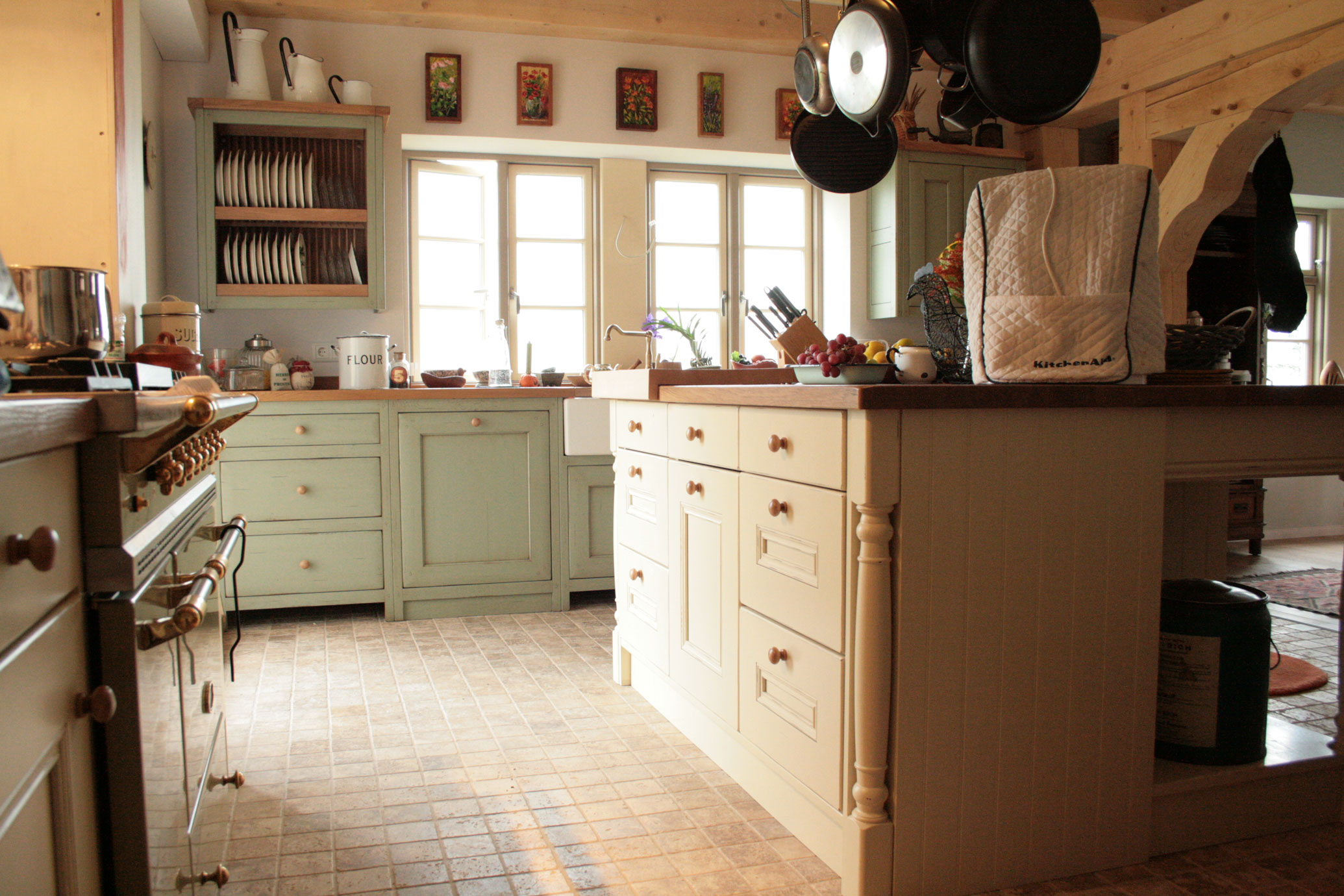 Landhausküche mit Kücheninsel, hellen Holzmöbeln und großzügigem Fenster, einladend und gemütlich eingerichtet Gemütliche Landhausküche mit Kücheninsel aus Holz, Hängeschränken, großen Fenstern und liebevollen Details wie Obstschale und Töpfen