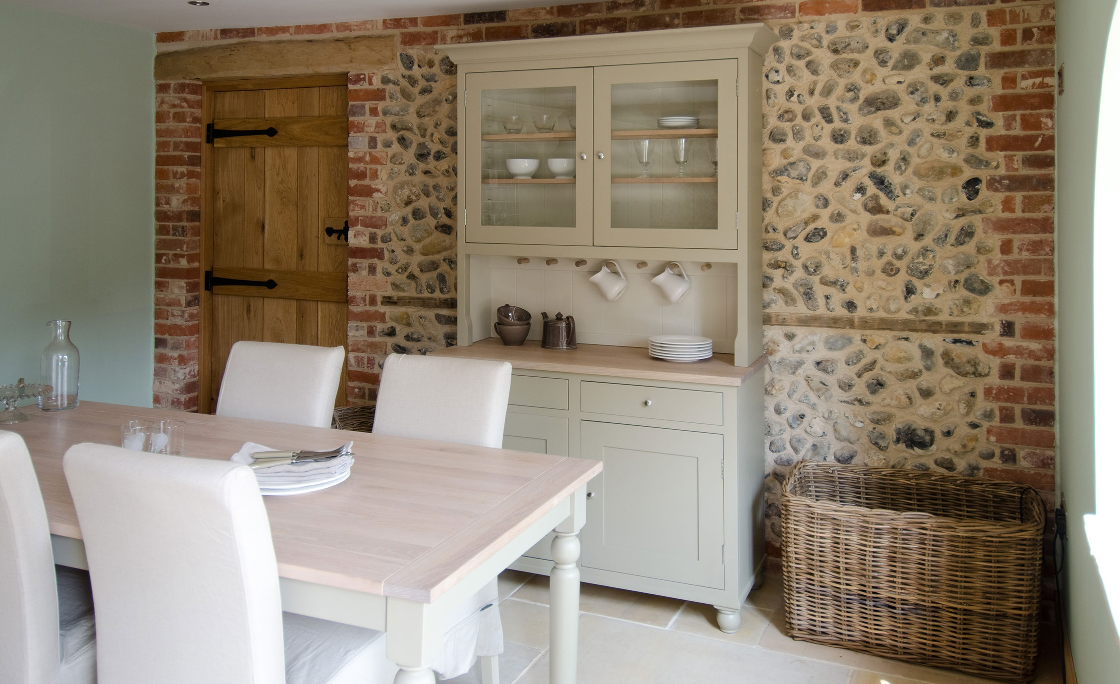 Esszimmer mit rustikalem Landhausstil, Holztisch und Vitrinenschrank vor gemauerter Steinwand Heller Essbereich mit massivem Holztisch, weißen Polsterstühlen und einem cremefarbenen Vitrinenschrank vor einer Wand aus Naturstein und Ziegeln, dazu ein Korb aus Naturgeflecht