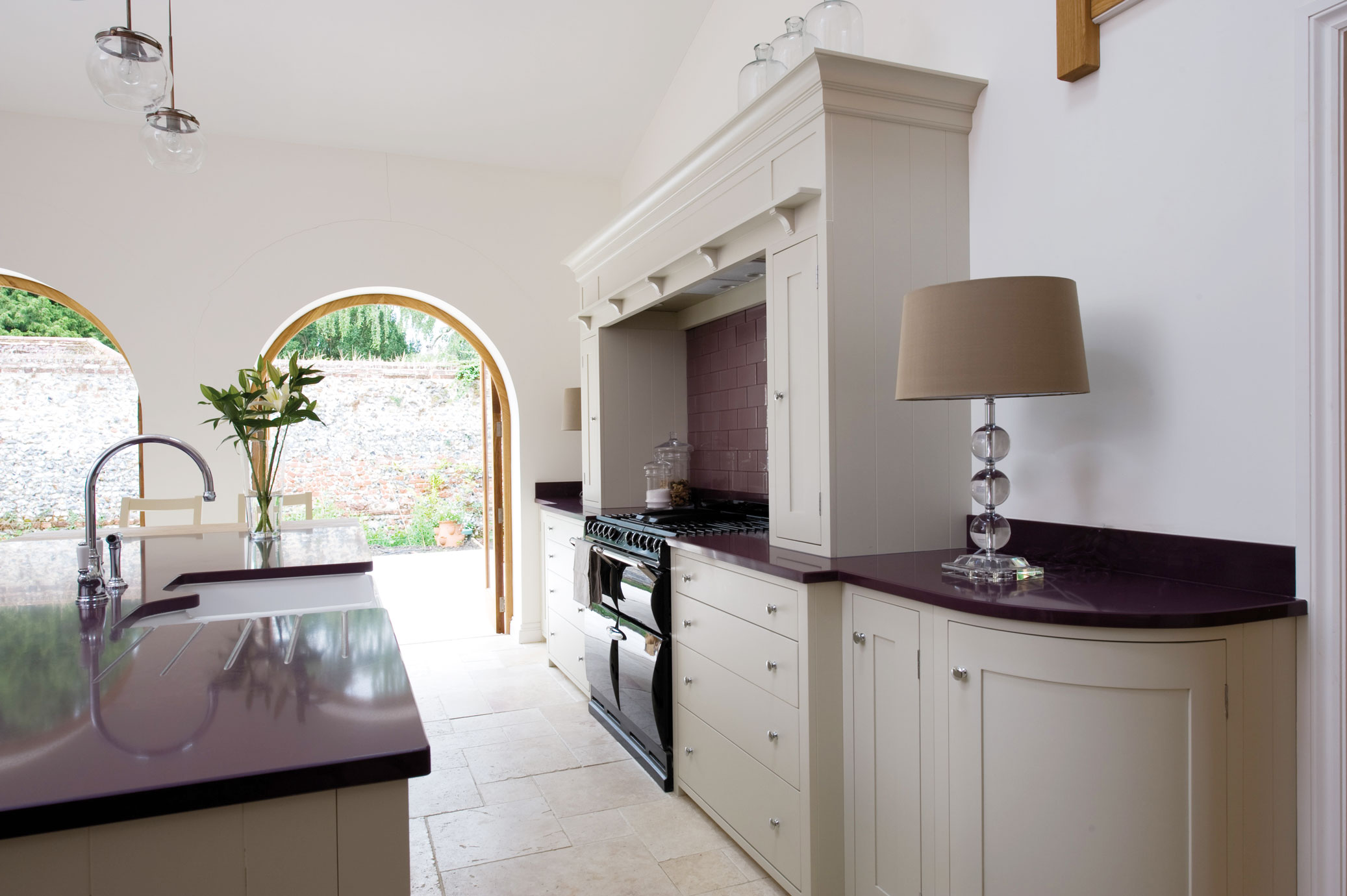 Moderne Landhausküche mit violetter Arbeitsplatte und eleganter Einrichtung Helle Landhausküche mit violetter Arbeitsplatte, cremefarbenen Schränken, Glaslampen und Blick auf den Garten durch große Rundbogenfenster