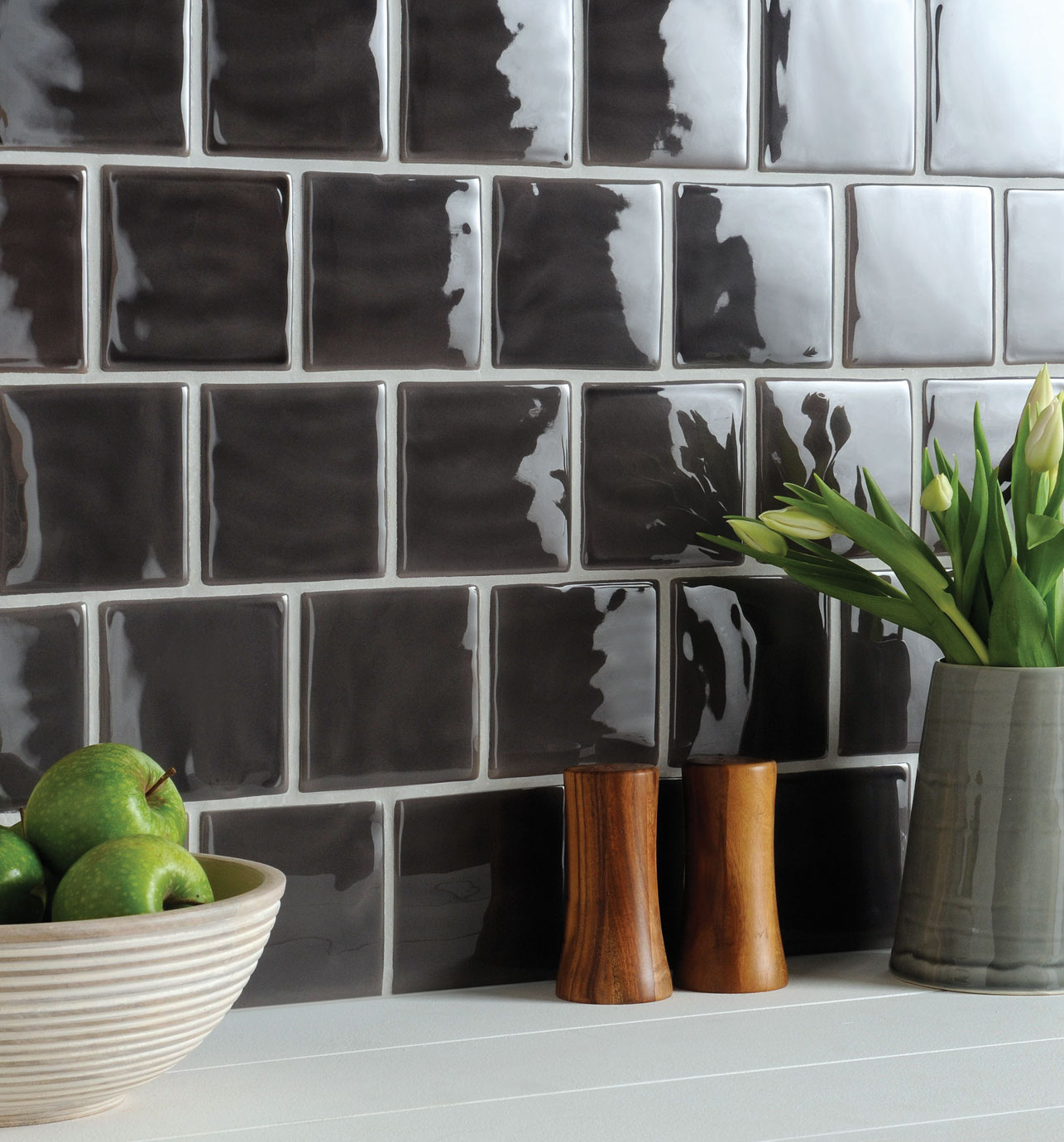 Glänzende dunkelgraue Wandfliesen in moderner Küche mit Schale grüner Äpfel und Tulpenvase als Dekoration Quadratische, dunkelgraue Fliesen mit glänzender Oberfläche an einer Küchenwand, davor eine Vase mit weißen Tulpen, zwei Holzgewürzmühlen und eine Schale mit grünen Äpfeln auf heller Arbeitsplatte
