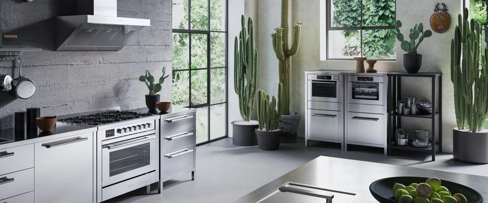 Moderne Loftküche: 90‑cm Gasherd aus Edelstahl mit großem Backofen, leistungsstarker Wand-Dunstabzug und passenden Einbauöfen; klare Fronten, Betonwand und hohe Fenster, dekoriert mit Kakteen