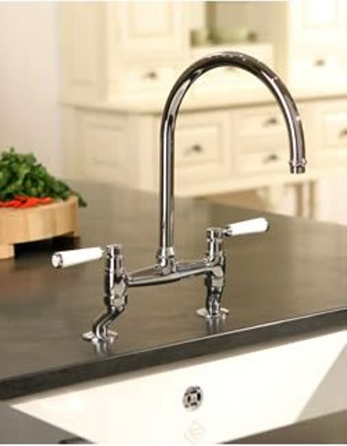 Elegante Küchenarmatur im Retro-Design mit zwei weißen Griffen auf moderner Kücheninsel Verchromte Küchenarmatur mit hohem Auslauf und weißen Griffen auf einer Kücheninsel, im Hintergrund helle Küchenschränke