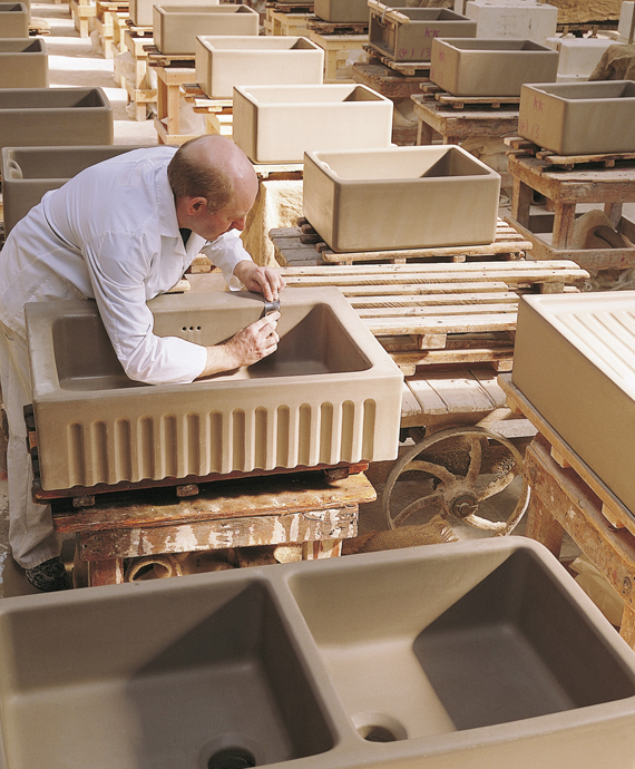 Handgefertigte Keramikspüle in traditioneller Manufaktur Ein Handwerker bearbeitet eine ungebrannte Keramikspüle in einer Werkstatt, umgeben von weiteren Spülen auf Holzpaletten