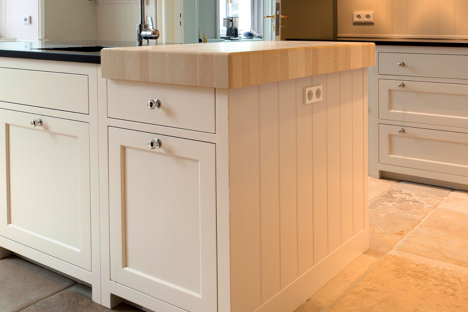 Detail einer cremefarbenen Kücheninsel im Landhausstil mit Massivholz-Arbeitsplatte, verchromten Knöpfen, Schubladen und seitlicher Doppelsteckdose auf Natursteinboden